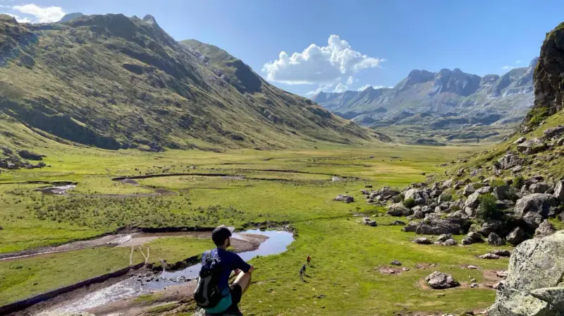 Un excursionista contempla un vasto valle de hecho, con un río serpenteante y montañas majestuosas bajo un cielo azul.