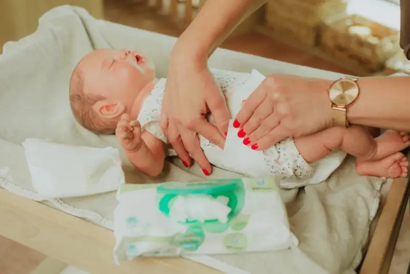 Delikatne dłonie z czerwonymi paznokciami pokazują, jak przewijać noworodka, używając chusteczek nawilżanych.