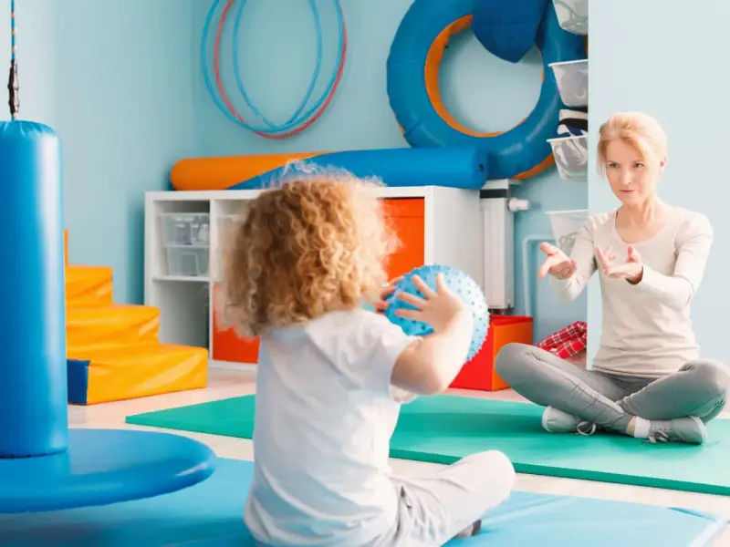 Ergotherapie für Kinder: Eine Therapeutin leitet ein Kind mit lockigem Haar an, das einen blauen Ball hält, in einem bunten Raum voller Spielgeräte.