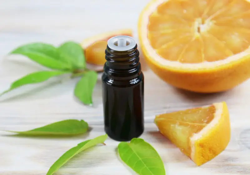 Flacon d'huile essentielle de citrus sinensis, entouré de feuilles vertes et de morceaux d'orange fraîche sur fond de bois clair.