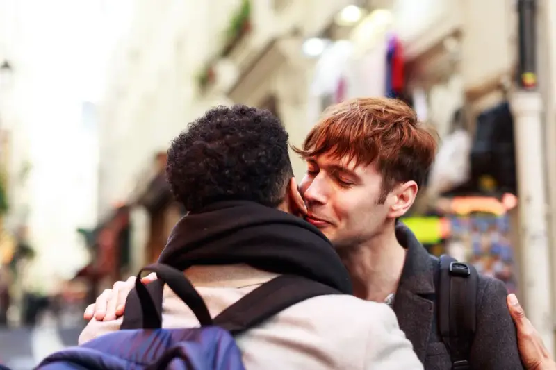 Un hombre joven con cabello rojizo susurra "bonjour" a su pareja, un momento íntimo en una calle parisina.