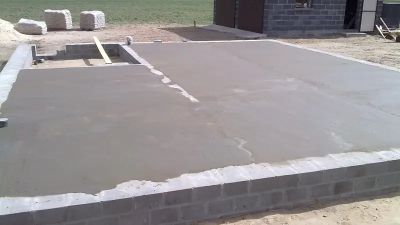 Świeżo wylany beton na chudziaka tworzy fundament pod budowę. W tle widać materiały budowlane i fragment budynku.