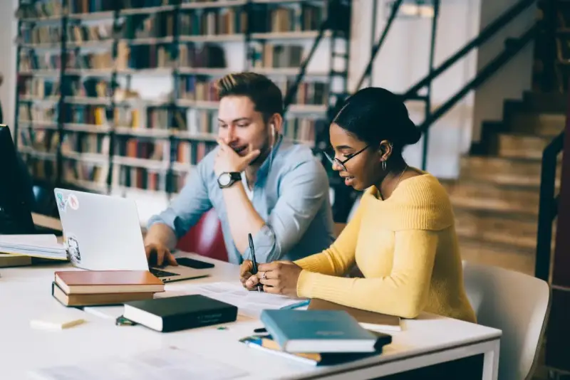 Studenti universitari collaborano a un progetto, immersi in libri e tecnologia.