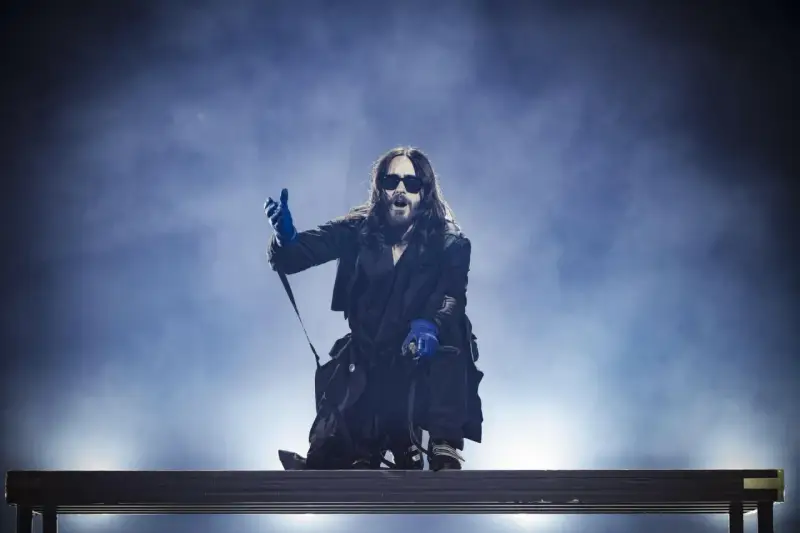 Jared Leto w niebieskich rękawiczkach, w okularach i długim płaszczu, klęczy na scenie. Jego zespół pewnie czeka na jego znak.