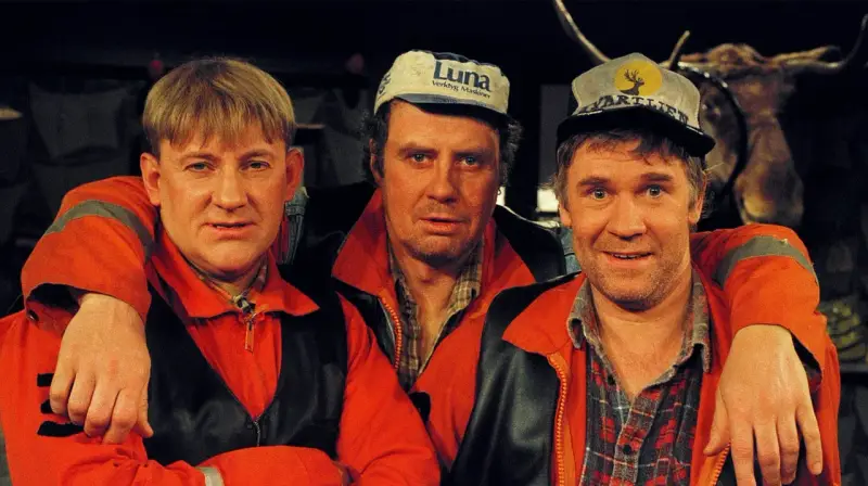 Tre män i orangea jackor och kepsar, en av dem med en uppstoppad älg i bakgrunden. Rollistan i Pistvakt.