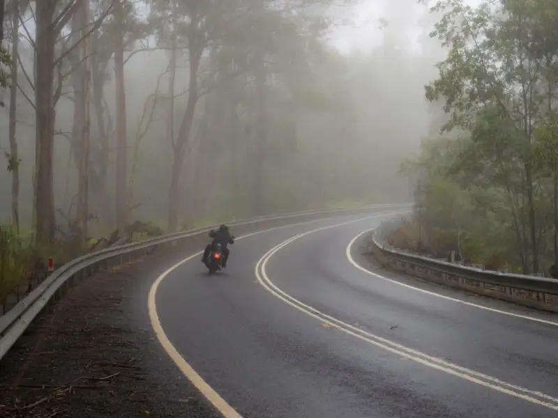 Motocyklista pokonuje zakręt na mokrej drodze, gdzie grozi mu aquaplaning. Mgła otula las, tworząc tajemniczą atmosferę.