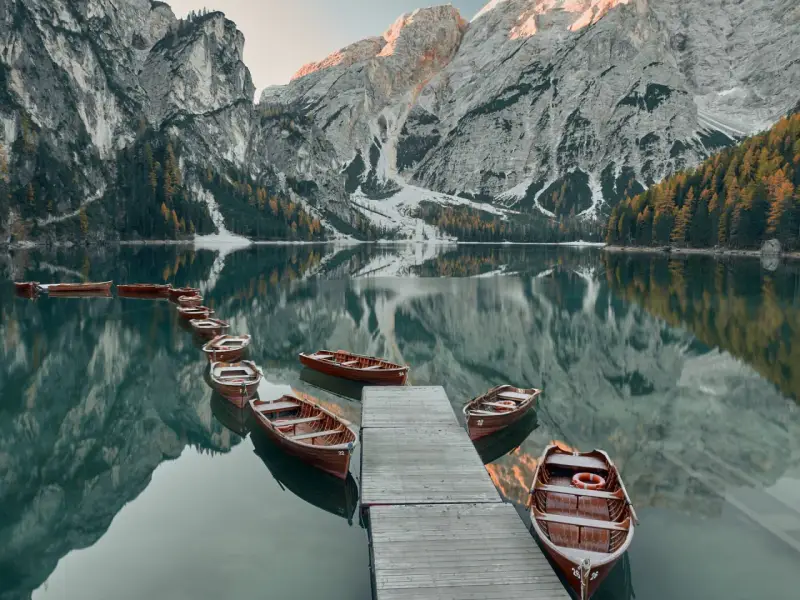 Drewniane łodzie przy drewnianym pomoście na spokojnym Lago di Braies, odbijające się w tafli wody, z majestatycznymi górami w tle.
