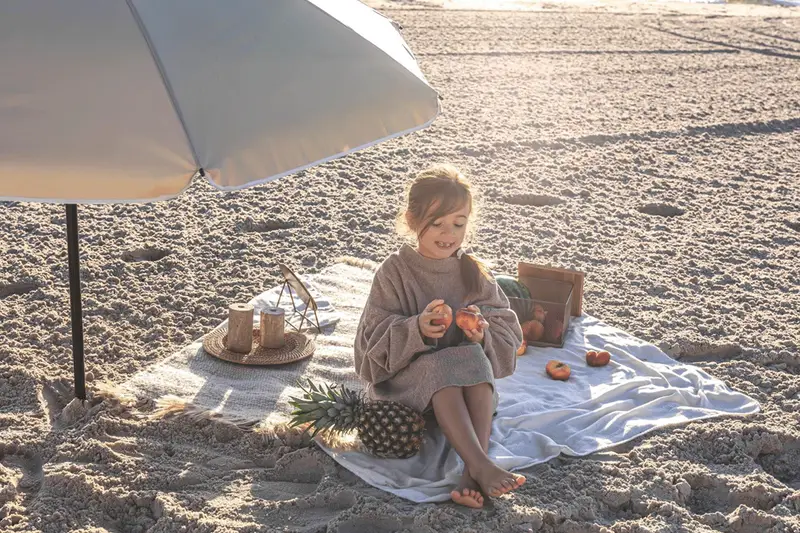 Dziewczynka na plaży pod parasolem, z owocami i ananasem. Idealne rzeczy, co zabrać na wakacje nad morze z dzieckiem.