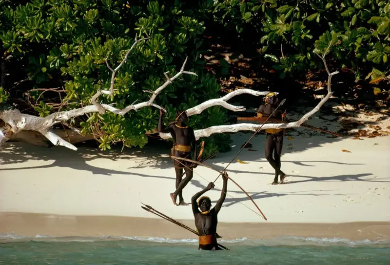 Trzech mężczyzn z plemienia Sentinelese, uzbrojonych w łuki i strzały, stoi na plaży. Sentinel Island, ich dom, chroni ich przed światem.