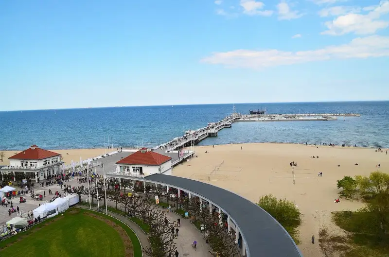 Sopot beach z molo, kawiarniami i spacerującymi ludźmi. W oddali widać statki.