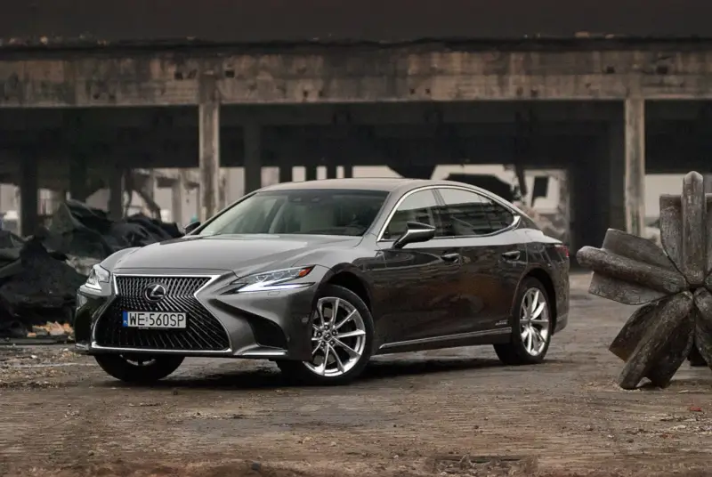 Szary Lexus LS 500h zaparkowany na tle zniszczonej budowli.