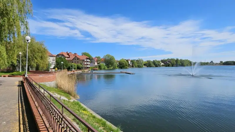 Spokojne jezioro zamkowe z fontanną i drewnianym pomostem, otoczone zielenią i zabudowaniami.