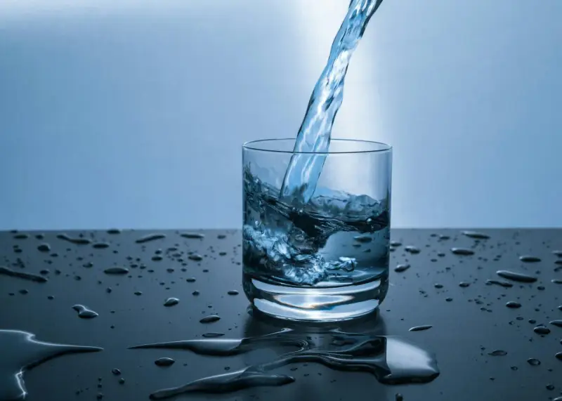 Un jet d'eau de source calcaire tombe dans un verre, créant des éclaboussures. Des gouttes d'eau parsèment la surface sombre.