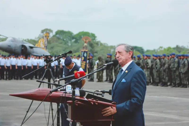 Elegancki garnitur to dobry wybór, gdy zastanawiasz się, jak się ubrać na przysięgę wojskową. Mężczyzna w granatowym garniturze przemawia na tle żołnierzy i samolotu.