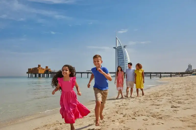 Rodzina cieszy się słonecznym dniem na plaży w Dubaju. Dzieci biegną w stronę morza, a w tle widać słynny hotel Burj Al Arab.