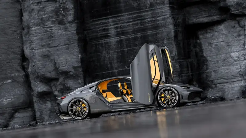 Szary Koenigsegg Gemera, czteromiejscny supersamochód z otwartymi drzwiami, prezentuje swoje żółte wnętrze na tle skalistej ściany.