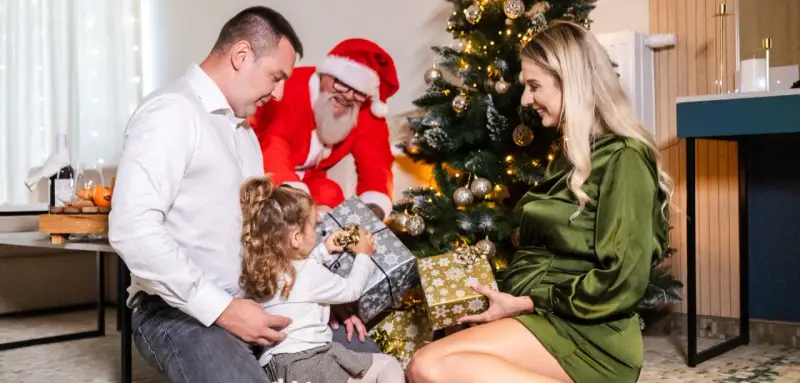 Rodzina otwiera prezenty przy choince, a Mikołaj uśmiecha się w tle. Wesołe święta w hotelu.