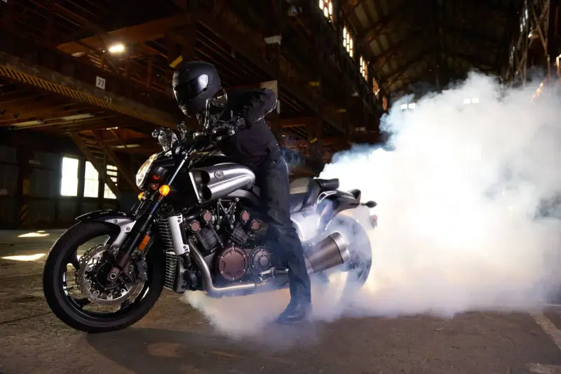 Motocyklista w kasku na Yamasze V-Max wykonuje burnout, tworząc kłęby dymu w opuszczonej hali.