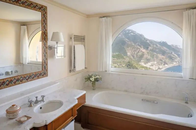 Un bagno di lusso con vasca, lavandino e specchio decorato, che offre una vista mozzafiato sulle montagne e sul mare. Tra i bagni più belli ed eleganti.