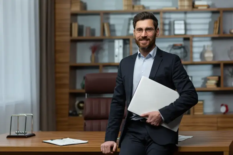 Uśmiechnięty dyrektor szkoły w garniturze, trzymający laptopa, stoi przy biurku w gabinecie.