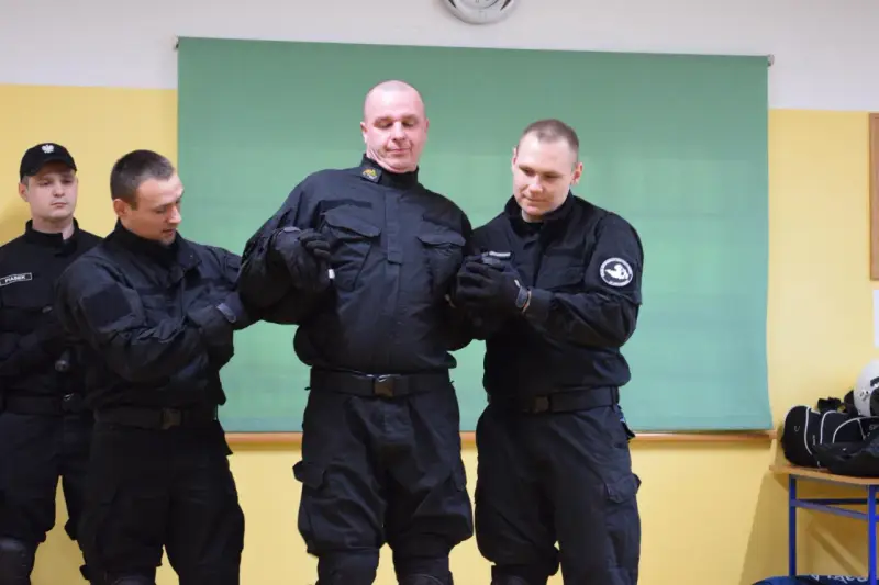 Funkcjonariusze w czarnych mundurach ćwiczą użycie środków przymusu kpk. Jeden z nich jest przytrzymywany przez dwóch kolegów.