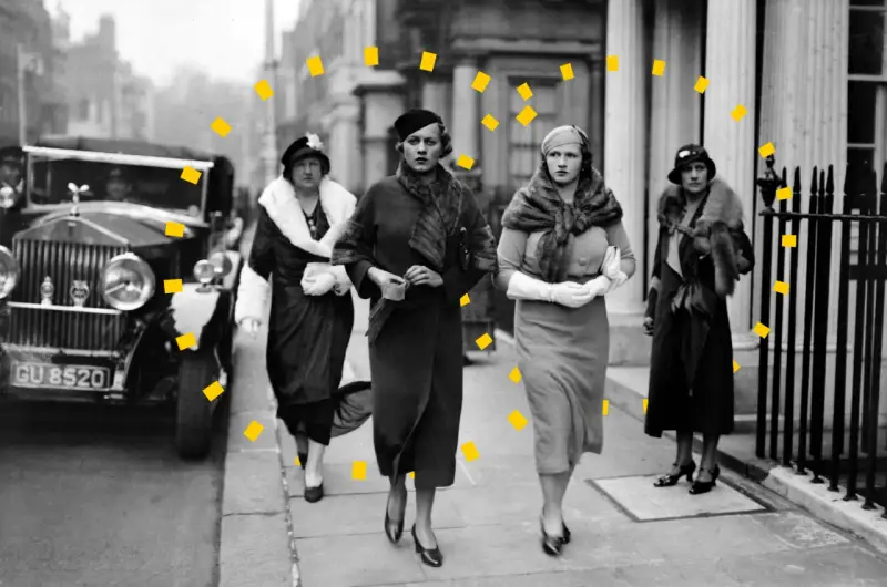 Vier Frauen in eleganter 30er Jahre Mode, Pelzmänteln und Hüten spazieren an einem Oldtimer entlang.