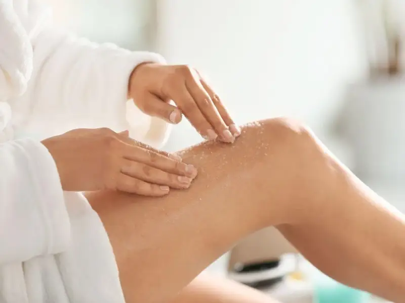 Kobieta w białym szlafroku wykonuje peeling nóg. To idealny peeling przed goleniem, by skóra była gładka.
