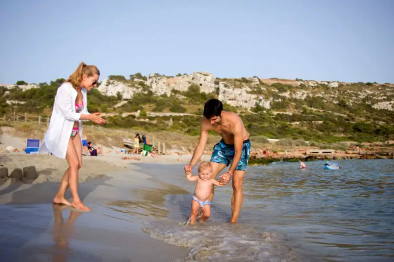 Familia disfrutando de un día en la playa en Menorca con niños. El padre ayuda a su bebé a dar sus primeros pasos en el agua, mientras la madre observa sonriente.