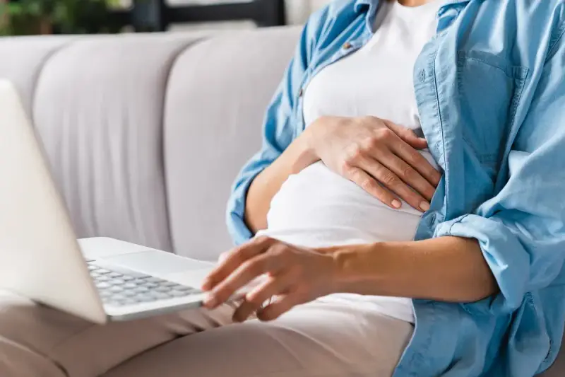 Kobieta w ciąży siedzi na kanapie, pisząc na laptopie wniosek o urlop macierzyński.