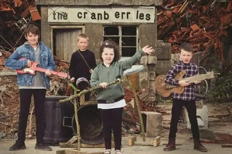 Quatre enfants jouent de la musique devant un panneau "the cranberries". L'un tient une guitare rose, un autre une guitare en bois.