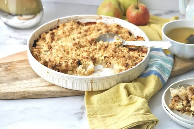 Ein goldbrauner Apfel-Crumble in einer Auflaufform, bereit zum Servieren. Daneben eine Tasse Tee und frische Äpfel.
