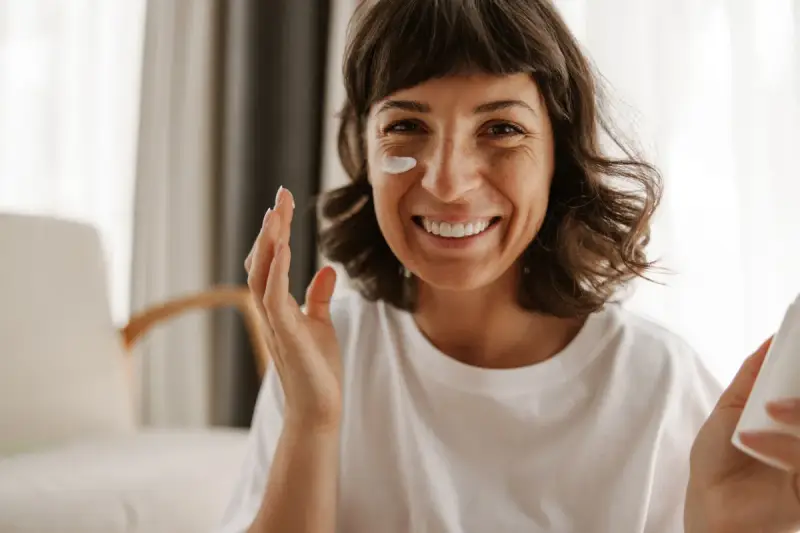 Mujer sonriente aplicándose crema en la mejilla, como si fueran vitaminas para el sol.