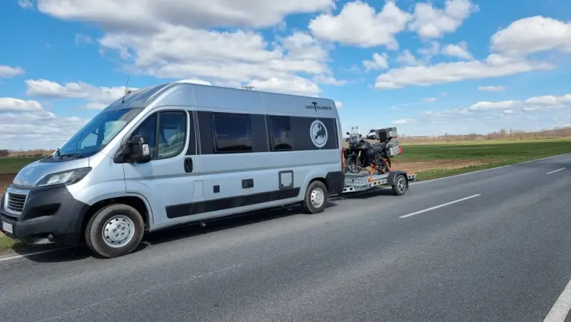Srebrny kamper z motocyklem na przyczepie jedzie drogą. Przygoda w Szwecji kamperem dopiero się zaczyna.