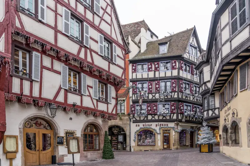 Fachwerkhäuser mit roten und weißen Fassaden in der Colmar Altstadt, geschmückt mit Lichterketten und Weihnachtsdekoration.