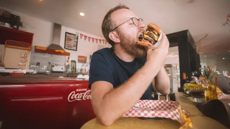 Mężczyzna z brodą i okularami zajada ogromnego burgera. W tle czerwona lodówka Coca-Coli, która może być jedną z ciekawostek o USA.