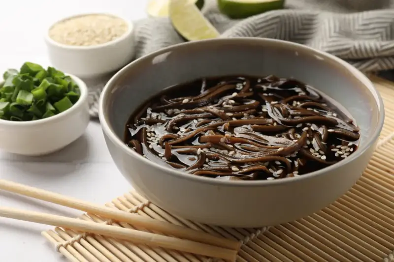 Miseczka z makaronem soba w sosie, posypana sezamem. Obok miseczki z posiekanym szczypiorkiem i sezamem. Zdrowy posiłek, mało makaron soba kcal.