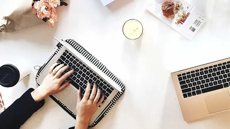 Kobieta pisze na laptopie, przygotowując wypowiedzenie umowy o dzieło. Obok świeca, muffinka i bukiet kwiatów.
