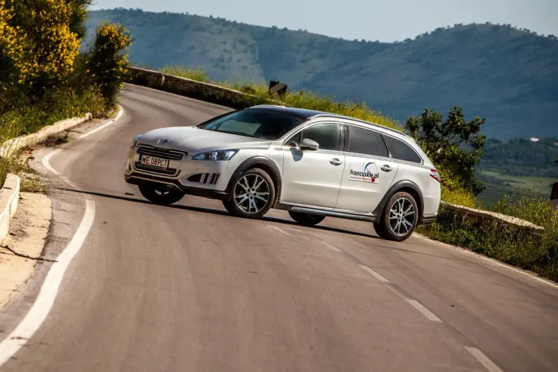 Białe kombi Peugeot 508 RXH na krętej drodze w górach.