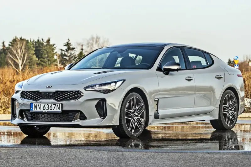 Elegancka Kia Stinger 2025 w kolorze srebrnym, zaparkowana na mokrej nawierzchni, z drzewami w tle.