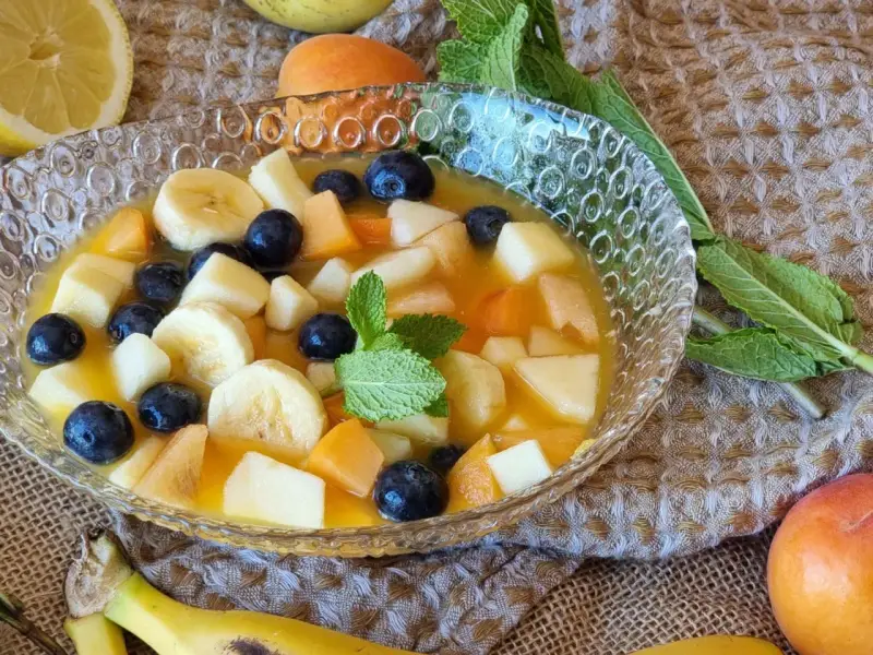 Deliciosa macedonia de frutas con plátano, arándanos y melocotón en un cuenco de cristal, decorada con menta.