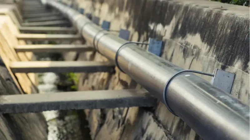 Metalowa rura biegnie wzdłuż betonowego koryta. To element kanalizacji ciśnieniowej, transportującej wodę.