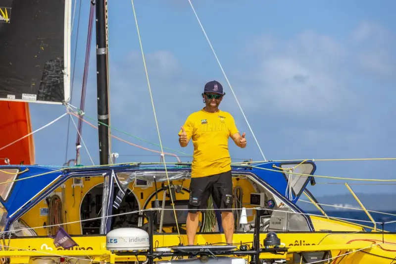 Arnaud Boissières, souriant, fait un double pouce levé sur son voilier jaune et bleu, prêt pour une nouvelle aventure en mer.