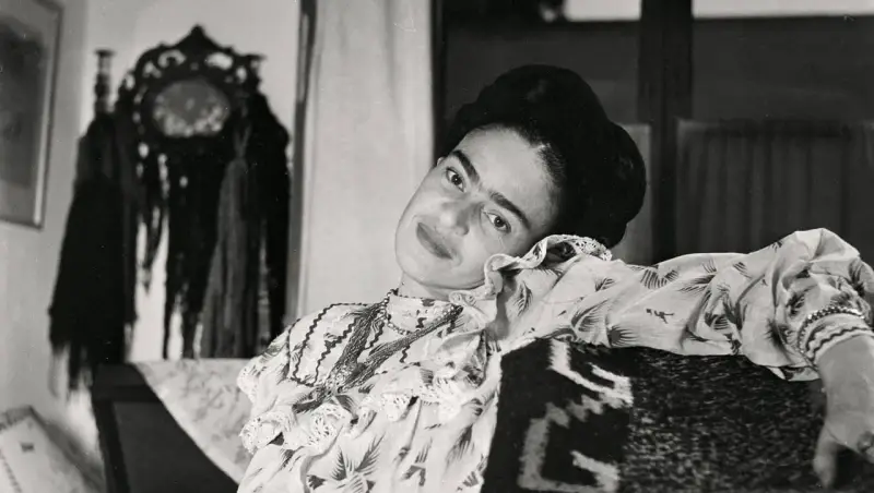 Frida Kahlo, le regard intense, pose dans une robe à volants. Derrière elle, un objet décoratif orné de franges, peut-être un indice sur son dernier tableau.