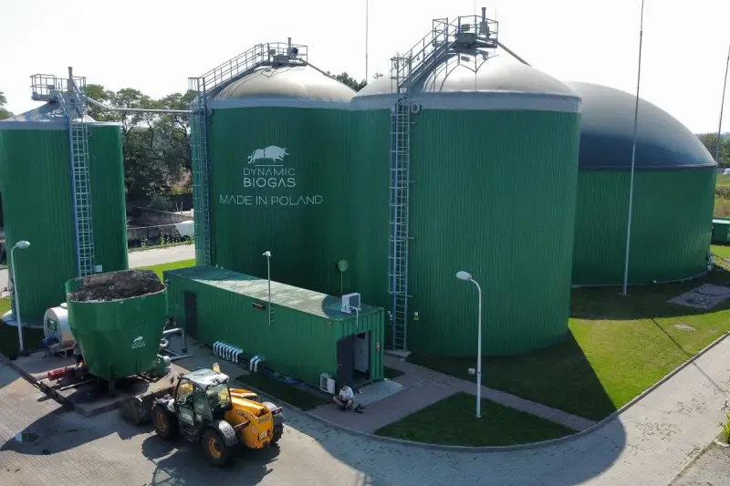 Zielone zbiorniki instalacji biogazowej, z logo "DYNAMIC BIOGAS MADE IN POLAND". Obok ładowarka i kontener.