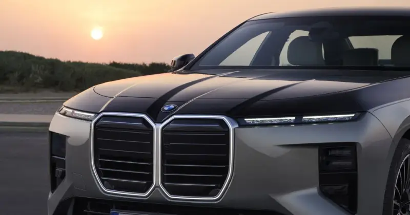 Przód luksusowego BMW M7 z podświetlaną atrapą chłodnicy i nowoczesnymi światłami, na tle zachodzącego słońca.