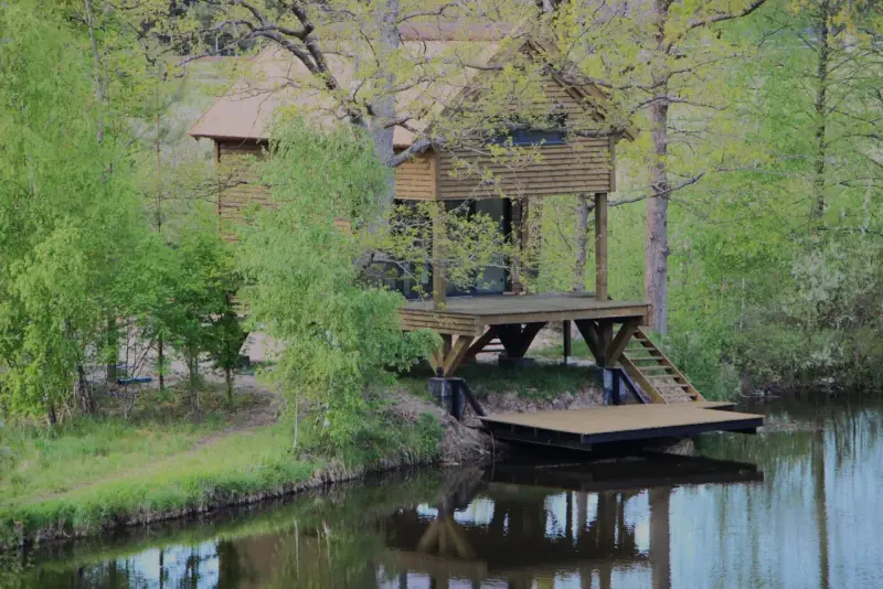 Drewniany domek na palach nad spokojnym jeziorem wiejskim, otoczony zielenią.