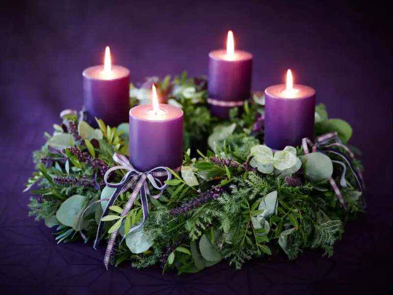 Four purple candles glow on a lush green Advent wreath, adorned with ribbons. The final candle's flame flickers, marking the 4th Advent.