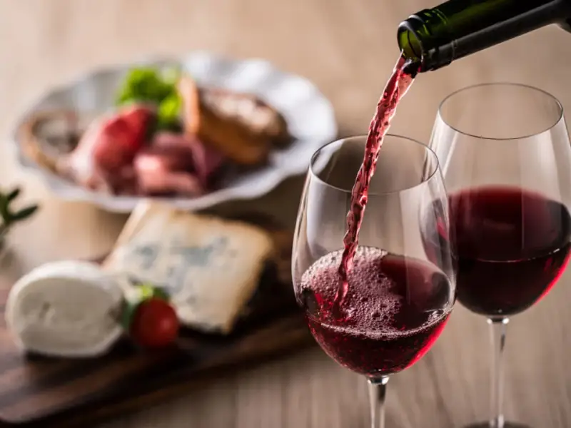 Rött vin hälls upp i glas, perfekt vin till grillat. Ost och charkuterier i bakgrunden.