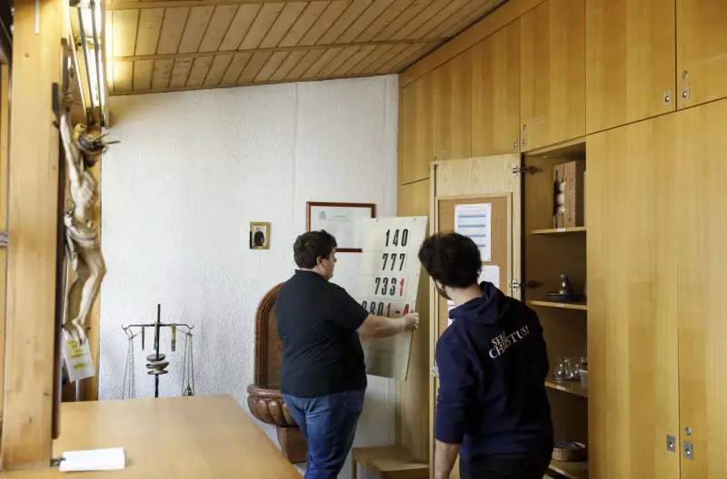 Zwei Männer im katholischen Priesterseminar hängen eine Liste mit Zahlen auf. Ein Kruzifix hängt an der Wand.