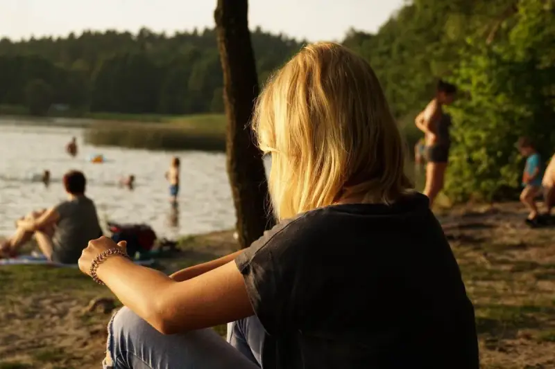 Dziewczyna z blond włosami siedzi nad jeziorem Trzechowskim, obserwując ludzi kąpiących się w wodzie.